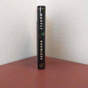 Puede incluir: Un libro negro de tapa dura con el t&iacute;tulo "Mostly Harmless" de Douglas Adams. La portada del libro presenta un extraterrestre verde con una cabeza grande y un cuerpo peque&ntilde;o.
