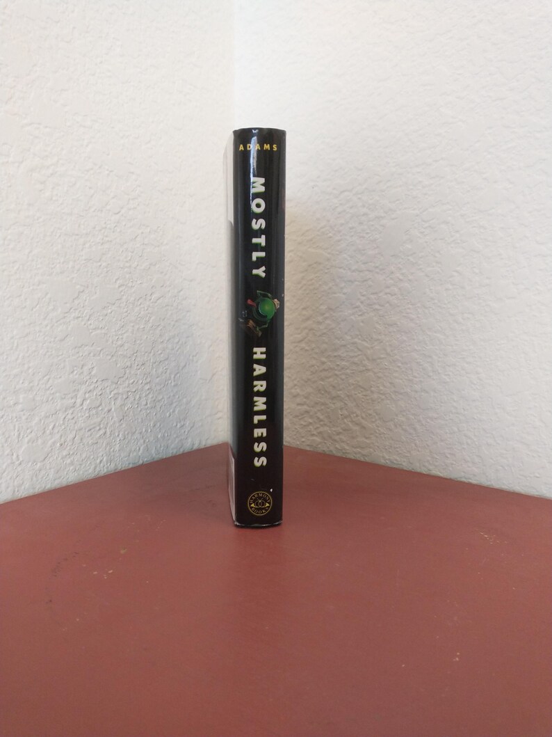 Puede incluir: Un libro negro de tapa dura con el t&iacute;tulo "Mostly Harmless" de Douglas Adams. La portada del libro presenta un extraterrestre verde con una cabeza grande y un cuerpo peque&ntilde;o.