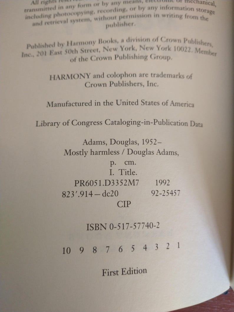 Puede incluir: Una portada de libro con el t&iacute;tulo "Mayormente inofensivo" de Douglas Adams. El libro forma parte de la serie "La gu&iacute;a del autoestopista gal&aacute;ctico". La portada es blanca con texto negro y una ilustraci&oacute;n en blanco y negro de una nave espacial.