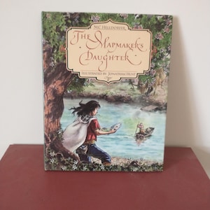 May include: The cover of a children's book titled "The Mapmaker's Daughter" by M.C. Helldorfer. The cover features an illustration of a young person in a red shirt and a white cape holding a map, standing on a riverbank. A small boat with two people is in the river.