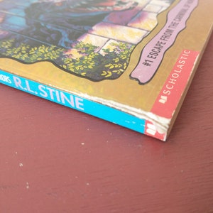 May include: A blue and gold paperback book with the title "Escape from the Carnival of Horrors" and the author "R.L. Stine". The book is part of the "Scholastic" series and is number 1 in the series.
