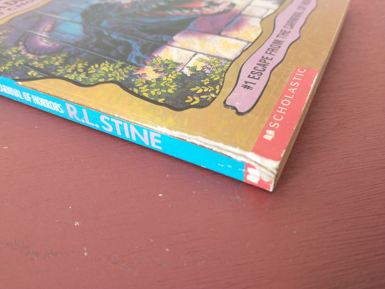 May include: A blue and gold paperback book with the title "Escape from the Carnival of Horrors" and the author "R.L. Stine". The book is part of the "Scholastic" series and is number 1 in the series.
