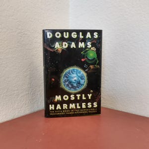 Puede incluir: Una portada de libro con fondo negro y un extraterrestre verde con la lengua fuera. El t&iacute;tulo del libro es "Mostly Harmless" y el autor es Douglas Adams. El subt&iacute;tulo es "The Fifth Book in the Increasingly Inaccurately Named Hitchhikers Trilogy."