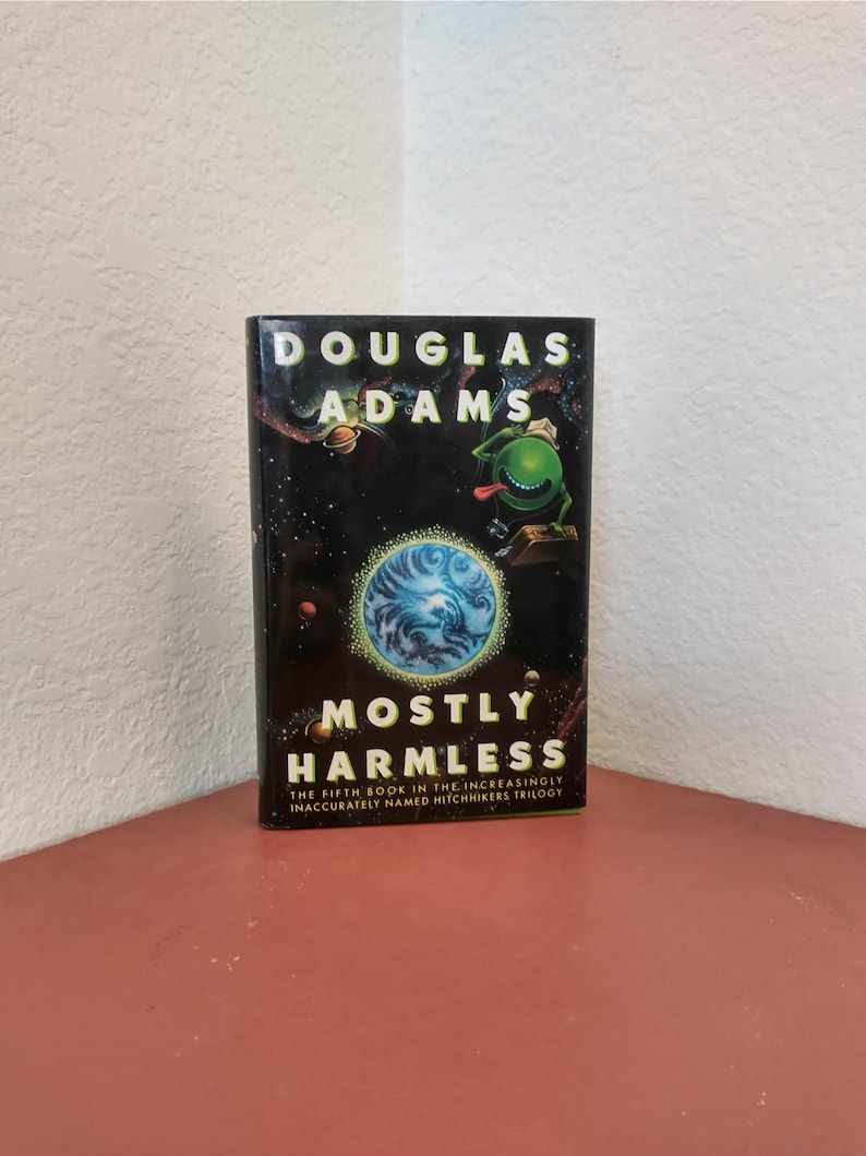 Puede incluir: Una portada de libro con fondo negro y un extraterrestre verde con la lengua fuera. El t&iacute;tulo del libro es "Mostly Harmless" y el autor es Douglas Adams. El subt&iacute;tulo es "The Fifth Book in the Increasingly Inaccurately Named Hitchhikers Trilogy."