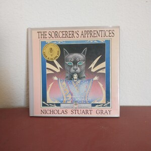 May include: A book cover with a pink background and a black cat with green eyes. The title of the book is "The Sorcerer's Apprentices" and the author is Nicholas Stuart Gray. The book is illustrated by Martin Springett.
