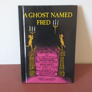 May include: A black book cover with a yellow title "A Ghost Named Fred" and a yellow illustration of two cherubs holding candlesticks. The book is by Nathaniel Benchley and illustrated by Ben Shecter. The book is an "I Can Read Book."