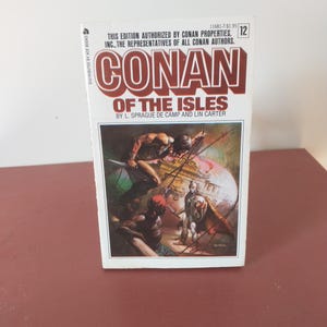 May include: A paperback book titled "CONAN OF THE ISLES" by L. Sprague de Camp and Lin Carter. The cover features a vibrant illustration of a battle scene with figures in action. The book is a vintage edition, with the text "DISTRIBUTED BY ACE BOOKS" visible.