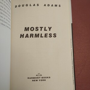 Puede incluir: Una cubierta de libro blanca con texto negro que dice "Douglas Adams" y "Mostly Harmless". La cubierta del libro tambi&eacute;n presenta un logotipo para Harmony Books, Nueva York.