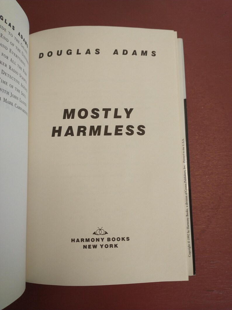 Puede incluir: Una cubierta de libro blanca con texto negro que dice "Douglas Adams" y "Mostly Harmless". La cubierta del libro tambi&eacute;n presenta un logotipo para Harmony Books, Nueva York.