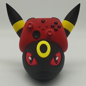 May include: A black and red 3D printed stand shaped like a Pokemon character with yellow accents. The stand is designed to hold an Xbox controller.