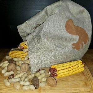 May include: A gray burlap sack with a brown squirrel design spills out corn cobs, peanuts, and walnuts onto a wooden cutting board. The corn is yellow with red kernels. The nuts are various shades of brown.