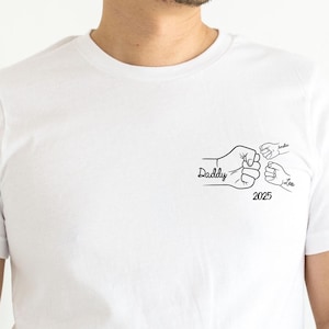 May include: A white short-sleeved t-shirt featuring a black line art graphic on the left chest. The design depicts a large fist bump with two smaller fists, accompanied by the text Daddy, 2025, and two names written in a cursive script font.
