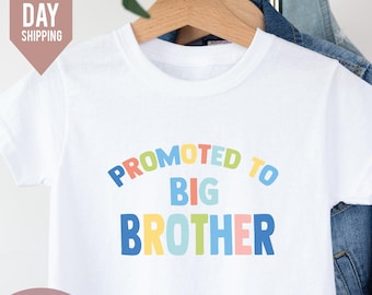 May include: White t-shirt with the text "Promoted to Big Brother" in rainbow colors. The text is in a curved arc.