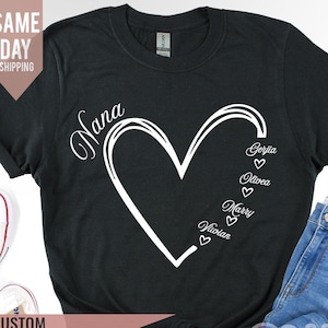 Puede incluir: Camiseta negra con un diseño de corazón blanco y la palabra "Nana" encima. Dentro del corazón están los nombres "Gertie", "Olivea", "Marry" y "Vivian" con un pequeño corazón al lado de cada nombre.