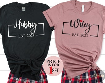 May include: A set of two black and pink t-shirts with the text "Hubby Est. 2023" and "Wifey Est. 2023" printed on them. The shirts are paired with red and black plaid pajama pants.