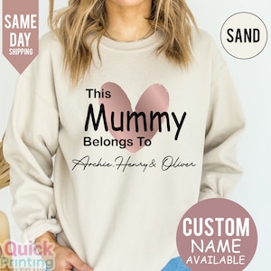 May include: Sand-colored sweatshirt with the text "This Mummy Belongs To" above the names Archie, Henry & Oliver, with a pink heart graphic. The sweatshirt also has a "Custom Name Available" graphic.