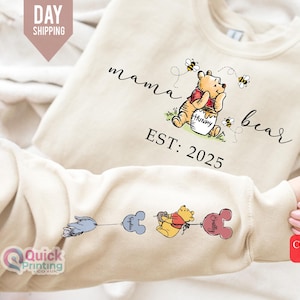 Könnte beinhalten: Ein beiger Pullover mit dem Text "mama bear est. 2025" und einem Cartoon-Bild von Winnie Puuh, der einen Honigtopf hält. Der Pullover hat auch ein kleines Cartoon-Bild von Winnie Puuh und einen Ballon am Ärmel.