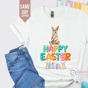 Op de afbeelding: Wit T-shirt met een kleurrijke "Happy Easter" afbeelding en een cartoon konijn.