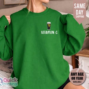 May include: A green sweatshirt featuring the text "VITAMIN G" and a graphic of a black and gold beverage. The sweatshirt is a solid green colour and has long sleeves. The design is printed on the front of the sweatshirt.