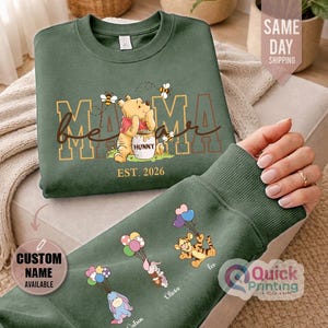 Puede incluir: Sudadera verde con Winnie the Pooh, el texto "Mama Bear" y "Hunny". La manga muestra personajes de dibujos animados con globos y nombres personalizados. El texto "Est. 2026" est&aacute; impreso debajo.
