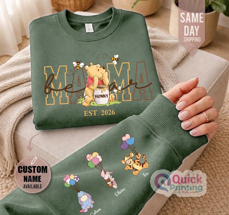 Puede incluir: Sudadera verde con Winnie the Pooh, el texto "Mama Bear" y "Hunny". La manga muestra personajes de dibujos animados con globos y nombres personalizados. El texto "Est. 2026" est&aacute; impreso debajo.