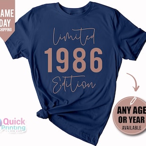 May include: A navy blue short-sleeved t-shirt featuring the text "Limited 1986 Edition" in a light pink color. The word "Limited" and "Edition" are written in a cursive script font, while the numbers "1986" are in a bold, block-style font.
