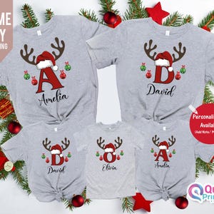 May include: A collection of gray Christmas t-shirts with reindeer antlers, Santa hats, and personalized letters and names. The shirts are decorated with red and green ornaments. The text "Amelia", "David", and "Olivia" are visible.