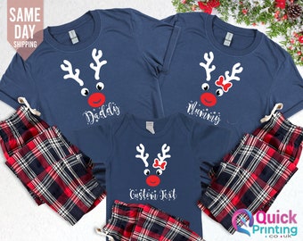 May include: A set of three navy blue t-shirts featuring a reindeer face design with the words "Daddy", "Mummy", and "Custom Text". The shirts are paired with red and black checked pyjama bottoms.