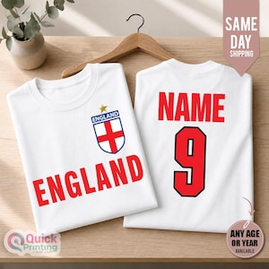 Könnte beinhalten: Zwei weiße T-Shirts mit rotem Text und Grafiken. Das vordere T-Shirt zeigt das Wort ENGLAND in Rot mit einem blau-weißen England-Wappen darüber. Das hintere T-Shirt zeigt das Wort NAME in Rot über der großen roten Ziffer 9.