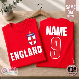 May include: Two red t-shirts displayed on a wooden hanger. The front of the left shirt features the word ENGLAND in white capital letters and an England crest with a gold star. The right shirt displays the word NAME and the number 9 in white.