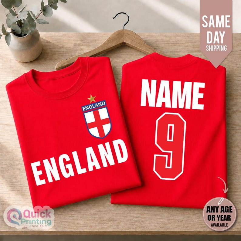May include: Two red t-shirts displayed on a wooden hanger. The front of the left shirt features the word ENGLAND in white capital letters and an England crest with a gold star. The right shirt displays the word NAME and the number 9 in white.