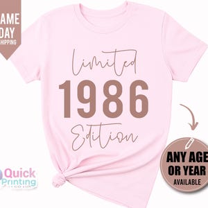 May include: A light pink short-sleeved t-shirt featuring the text Limited 1986 Edition in a brown font. The shirt has a crew neckline and a knotted hem detail on the lower left side. The design is centered on the front of the garment.