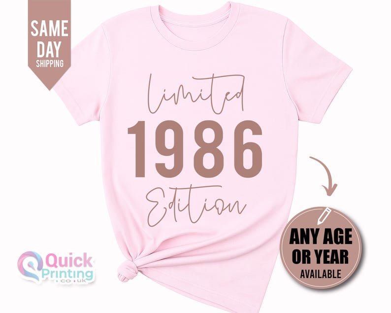 May include: A light pink short-sleeved t-shirt featuring the text Limited 1986 Edition in a brown font. The shirt has a crew neckline and a knotted hem detail on the lower left side. The design is centered on the front of the garment.