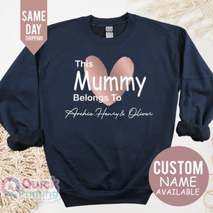 May include: Navy blue sweatshirt with the text "This Mummy Belongs To" and the names Archie, Henry & Oliver. A rose gold heart shape is above the word "Mummy". The sweatshirt also has a "Custom Name Available" option.