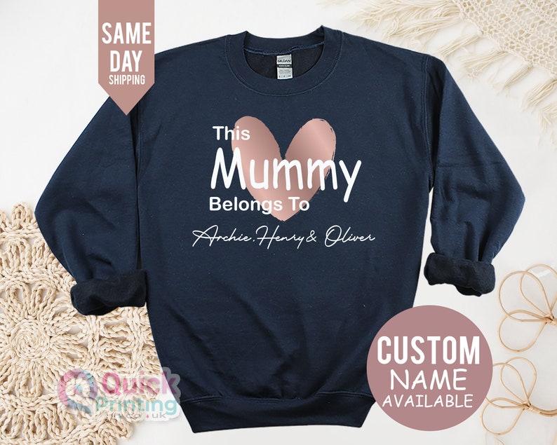 May include: Navy blue sweatshirt with the text "This Mummy Belongs To" and the names Archie, Henry & Oliver. A rose gold heart shape is above the word "Mummy". The sweatshirt also has a "Custom Name Available" option.