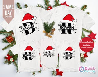 Pode incluir: Camisetas e body de Natal brancos para a família. Cada camisa apresenta um design de chapéu de Papai Noel com uma letra e um nome. O texto "SAME DAY SHIPPING" é visível. Um círculo vermelho diz "Personalisation Available".