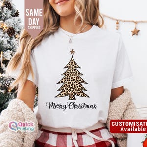 May include: White t-shirt with a Christmas tree design in a leopard print pattern. The tree is topped with a star, and the words "Merry Christmas" are written below in a script font. The shirt also has the text "Customisation Available".