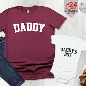 Può includere: Maglietta color borgogna con la parola "DADDY" in lettere bianche e un body bianco con il testo "DADDY'S BOY" in lettere nere.