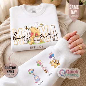 Puede incluir: Sudadera blanca con dise&ntilde;o de Winnie the Pooh, texto "Mama Bear" y "Est. 2026". La manga muestra personajes de dibujos animados con globos y nombres personalizados. La sudadera tiene cuello redondo y mangas largas. Tambi&eacute;n incluye el texto "Custom Name Available".