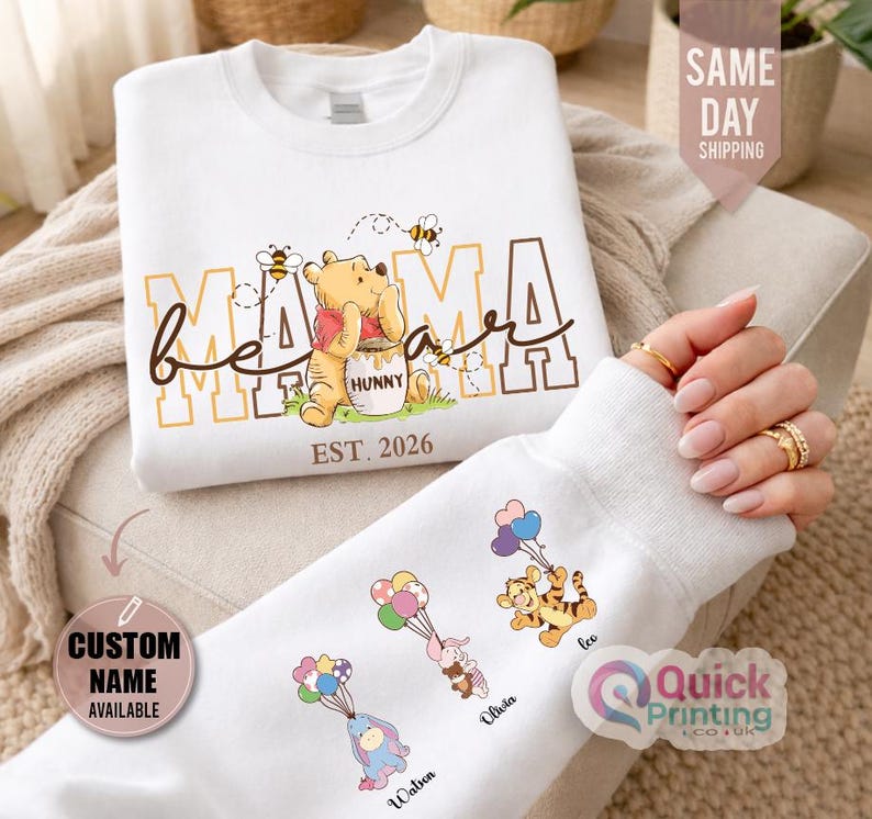 Puede incluir: Sudadera blanca con dise&ntilde;o de Winnie the Pooh, texto "Mama Bear" y "Est. 2026". La manga muestra personajes de dibujos animados con globos y nombres personalizados. La sudadera tiene cuello redondo y mangas largas. Tambi&eacute;n incluye el texto "Custom Name Available".
