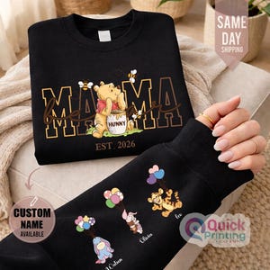 Puede incluir: Sudadera negra con dise&ntilde;o dorado de "MAMA" y Winnie the Pooh. La manga muestra personajes de dibujos animados con globos. El texto "EST. 2026" est&aacute; presente. El texto "CUSTOM NAME AVAILABLE" tambi&eacute;n es visible.