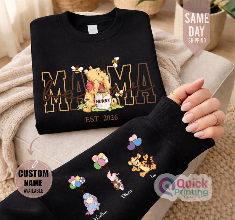 Puede incluir: Sudadera negra con dise&ntilde;o dorado de "MAMA" y Winnie the Pooh. La manga muestra personajes de dibujos animados con globos. El texto "EST. 2026" est&aacute; presente. El texto "CUSTOM NAME AVAILABLE" tambi&eacute;n es visible.