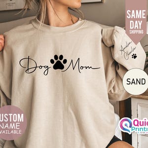 May include: A beige sweatshirt with the text "Dog Mom" in black script with a black paw print between the words. The sweatshirt has a ruffled detail on the sleeves. The text "Custom Name Available" is in a white box on the bottom left of the image. The text "Sand" is in a white circle on the bottom right of the image.