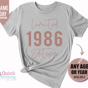 May include: A light gray short-sleeve t-shirt featuring the text Limited 1986 Edition in a muted rose color. The word Limited and Edition are written in a cursive script font, while the number 1986 is displayed in a large, bold, sans-serif font.