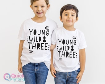 May include: Two white t-shirts with black text that reads "Young Wild & Three" on each shirt. The shirts are being worn by two children.