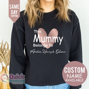 May include: Black sweatshirt with the text "This Mummy Belongs To Archie, Henry & Oliver" and a pink heart graphic. Additional text includes "Custom Name Available" and "Same Day Shipping".