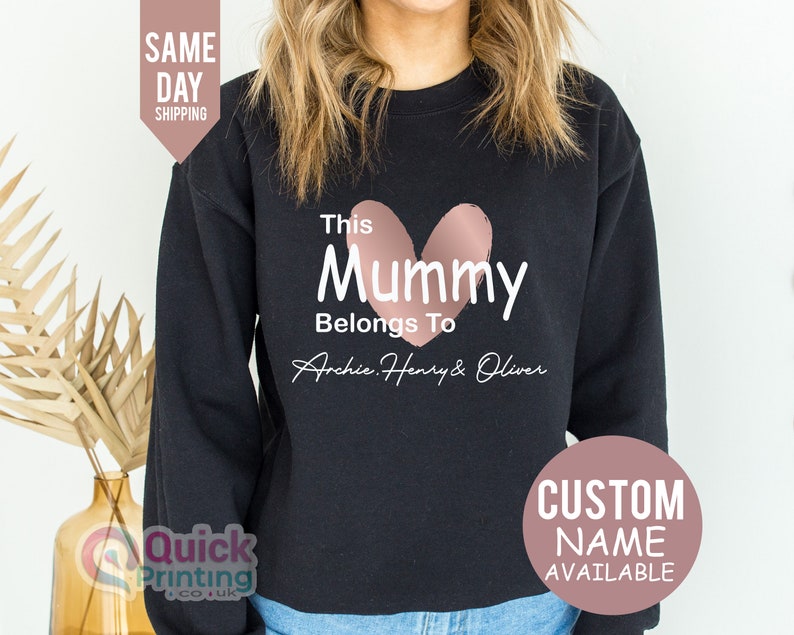 May include: Black sweatshirt with the text "This Mummy Belongs To Archie, Henry & Oliver" and a pink heart graphic. Additional text includes "Custom Name Available" and "Same Day Shipping".