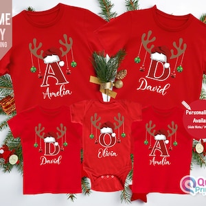 May include: Red Christmas-themed t-shirts and a onesie with reindeer antlers, Santa hats, and personalized names. The shirts and onesie have the names Amelia, David, and Olivia. The text "Personalisation Available" is also visible.