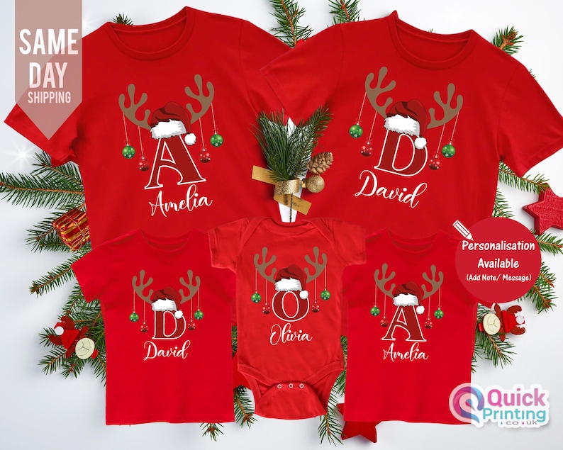 May include: Red Christmas-themed t-shirts and a onesie with reindeer antlers, Santa hats, and personalized names. The shirts and onesie have the names Amelia, David, and Olivia. The text "Personalisation Available" is also visible.