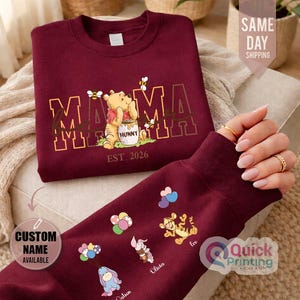 Puede incluir: Sudadera burdeos con un dise&ntilde;o de Winnie the Pooh, la palabra "MAMA" y "EST. 2026". La manga muestra personajes de dibujos animados con nombres. Tambi&eacute;n es visible el texto "CUSTOM NAME AVAILABLE".